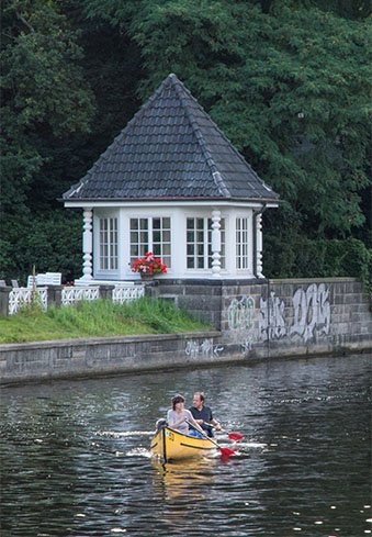 Canoeists paddle past a riverside hut.